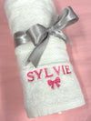 Rolled white baby towel tied with a grey satin ribbon, embroidered with the name Sylvie in pink and a pink bow