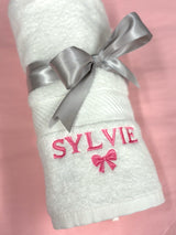 Rolled white baby towel tied with a grey satin ribbon, embroidered with the name Sylvie in pink and a pink bow
