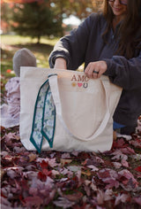 Custom embroidered natural tote with AMC initials, sun, heart and horseshoe icons and a scarf tied on the handle